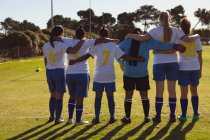 Visão traseira de diversas jogadoras de futebol do sexo feminino de pé com braço ao redor no campo esportivo — Fotografia de Stock