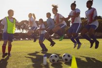 Vista laterale di allenatore caucasico aiutare diversi giocatori di calcio femminile con esercizio di salto sul campo sportivo — Foto stock