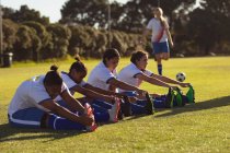 Vista lateral de diversas jugadoras de fútbol haciendo ejercicio de estiramiento en el campo en un día soleado - foto de stock
