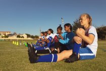 Vista lateral de diversas jogadoras de futebol do sexo feminino fazendo exercício de alongamento de perna no campo em um dia ensolarado — Fotografia de Stock