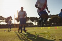 Низкий угол обзора различных женщин-футболистов, прыгающих через препятствия во время тренировок на спортивной площадке в солнечный день — стоковое фото