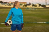 Vista frontal do pensativo jogador de futebol feminino caucasiano de pé com futebol no campo de esportes — Fotografia de Stock