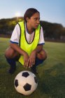Primer plano de la reflexiva jugadora de fútbol afroamericana agachada con el fútbol en el campo de deportes - foto de stock