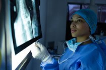 Vista lateral de la hermosa joven cirujana de raza mixta examinando rayos X en la caja de luz en el quirófano del hospital. Cirujano lleva bata quirúrgica, máscara, gorra y guantes . - foto de stock