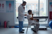 Seitenansicht des kaukasischen männlichen Arztes, der einen älteren Mischlingspatienten mit Stethoskop im Krankenhaus untersucht — Stockfoto