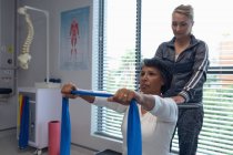 Visão lateral de fisioterapeuta feminina caucasiana dando fisioterapia a paciente mestiça com banda de resistência no hospital — Fotografia de Stock