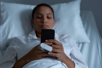 Вид спереду красива змішана раса жінка-пацієнтка, що використовує мобільний телефон, лежачи на ліжку в палаті в лікарні — стокове фото