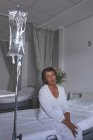 Vista frontal de una paciente de raza mixta sentada en la cama con IV de pie frente a ella en la sala del hospital - foto de stock
