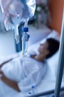 Primer plano del goteo intravenoso con paciente de raza mixta acostada en la cama en el fondo de la sala en el hospital - foto de stock
