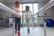 Vorderansicht einer kaukasischen Physiotherapeutin, die einer Patientin hilft, mit parallelen Stangen im Krankenhaus zu gehen — Stockfoto