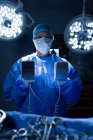 Portrait d'une chirurgienne métisse tenant un défibrillateur alors qu'elle regardait une caméra en salle d'opération à l'hôpital — Photo de stock
