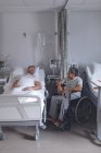 Seitenansicht diverser männlicher Patienten, die auf der Station im Krankenhaus miteinander interagieren. Kaukasischer Patient liegt im Bett, während Mischlingspatient im Rollstuhl sitzt. — Stockfoto