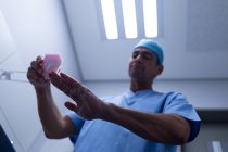 Low angle view of handsome middle aged Caucasian male surgeon scrubbing hands with brush and soap at sink in hospital. He is wearing surgical gown and cap. — Stock Photo