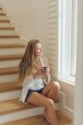 Side view of happy Caucasian woman with coffee cup looking through window while sitting on stairs at home — Stock Photo
