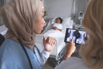 Primer plano de diversas doctoras discutiendo sobre el informe de rayos X en tabletas digitales en la sala del hospital. En el fondo, la paciente de raza mixta está durmiendo en la cama en la sala del hospital. . - foto de stock