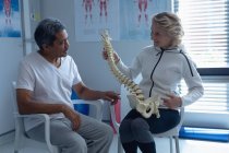 Vorderseite der kaukasischen Physiotherapeutin erklärt männlichen Patienten im Krankenhaus das Wirbelsäulenmodell — Stockfoto