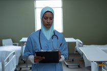 Visão frontal de mestiça médica feminina em hijab escrevendo em prancheta na enfermaria do hospital — Fotografia de Stock