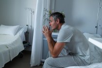 Vue latérale du patient masculin caucasien réfléchi assis sur le lit avec les coudes sur le genou dans la salle à l'hôpital — Photo de stock