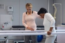 Вид збоку кавказької жінки фізіотерапевт допомагаючи змішаної раси жіночого пацієнта ходити з паралельними барами в лікарні — стокове фото