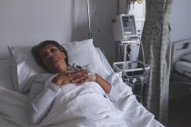 Vue en angle élevé d'une patiente métissée réfléchie se relaxant au lit dans le service de l'hôpital. Femme garde les mains sur la poitrine . — Photo de stock