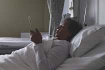 Vista lateral da bela paciente mestiça madura usando tablet digital na enfermaria do hospital . — Fotografia de Stock