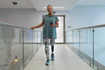 Vista frontal de una paciente caucásica discapacitada con una pierna protésica usando teléfono móvil mientras camina por el pasillo en el hospital - foto de stock