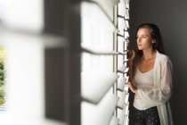 Вид вдумливого кавказька жінка дивиться через вікно сліпих в домашніх умовах — стокове фото
