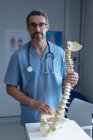 Retrato de cirujano ortopédico masculino caucásico con estetoscopio alrededor del cuello sosteniendo el modelo de columna vertebral en el hospital - foto de stock