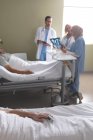 Visão lateral do paciente caucasiano do sexo masculino relaxando na cama enquanto diversos médicos interagem uns com os outros na enfermaria do hospital. Em primeiro plano paciente caucasiano do sexo masculino está dormindo na cama . — Fotografia de Stock
