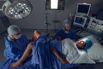 Vista de ángulo alto del cirujano femenino caucásico que examina a la mujer embarazada durante el parto mientras el hombre sostiene su mano en el quirófano en el hospital - foto de stock