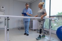 Front view of Caucasian male physiotherapist writing on clipboard and Caucasian female amputee patient walking with parallel bars in the hospital — Stock Photo