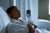 Вид збоку вдумливої змішаної раси пацієнтка-жінка лежить в ліжку з руками на грудях в палаті в лікарні. Порожнє ліжко і монітор видно у фоновому режимі . — стокове фото