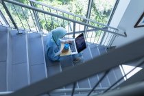 Vista alta del médico femenino de raza mixta en hijab usando computadora portátil en la escalera en el hospital - foto de stock