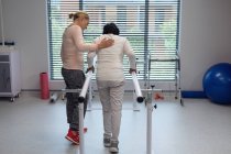 Visão traseira da fisioterapeuta feminina caucasiana ajudando a paciente mestiça a caminhar com barras paralelas no hospital — Fotografia de Stock