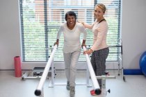 Visão frontal da fisioterapeuta feminina caucasiana ajudando a paciente a caminhar com barras paralelas no hospital — Fotografia de Stock