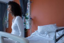 Vue latérale d'une patiente métisse réfléchie assise sur le lit tout en regardant loin de la caméra à l'hôpital — Photo de stock