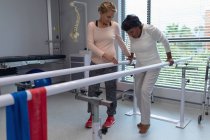 Вид сбоку кавказской женщины-физиотерапевта, помогающей пациентке смешанной расы ходить с параллельными решетками в больнице — стоковое фото