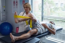 Vue latérale du médecin féminin caucasien examinant la jambe du patient âgé métis avec bande de résistance à l'hôpital — Photo de stock