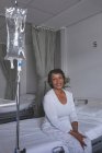 Retrato de una hermosa paciente de raza mixta sentada en la cama mientras sonríe hacia la cámara en la sala del hospital. IV stand está al lado de la cama
. - foto de stock