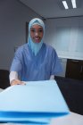 Vista frontale di chirurgo di razza mista in hijab in piedi con file di referto medico alla reception in ospedale . — Foto stock