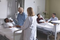 Вид сбоку различных женщин-врачей, взаимодействующих с пациенткой в палате больницы. На заднем плане кавказский мужчина держит за руку азиатку, лежащую в постели в больнице . — стоковое фото