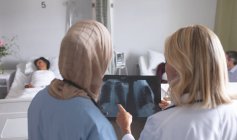 Rear view of diverse female doctors discussing x-ray report in the ward at hospital. In the background diverse patients are sleeping in bed in the ward in hospital. — Stock Photo