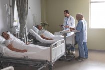 Побочный обзор различных врачей, взаимодействующих со старшим пациентом смешанной расы, в то время как белый мужчина спит рядом с ним в палате больницы — стоковое фото