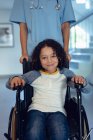 Retrato de niño discapacitado de raza mixta en silla de ruedas con doctora en el pasillo del hospital - foto de stock