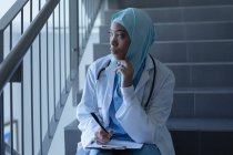 Vorderansicht einer nachdenklichen gemischtrassigen Ärztin im Hijab, die auf der Treppe sitzt und Stift und Klemmbrett im Krankenhaus hält. — Stockfoto