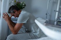 Боковий вид стурбований Кавказького чоловічого пацієнта сидячи на ліжку з лікті на коліні в палаті в лікарні — стокове фото
