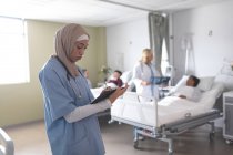 Vista lateral de una hermosa doctora de raza mixta en hiyab escribiendo en el portapapeles en la sala del hospital. En el fondo diversos médicos están interactuando con sus pacientes
. - foto de stock