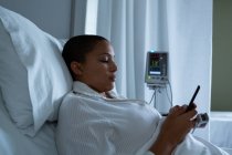 Vista lateral del hermoso paciente femenino de raza mixta usando teléfono móvil mientras está acostado en la cama en la sala en el hospital - foto de stock