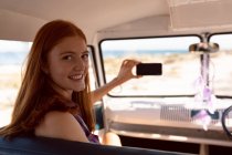 Vista frontal de la hermosa mujer caucásica mirando a la cámara mientras toma selfie con teléfono móvil en caravana en la playa - foto de stock