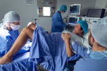 Вид збоку різних хірургів вивчення вагітної жінки під час пологів, а Кавказький чоловік тримає її руку в операційній кімнаті в лікарні — стокове фото